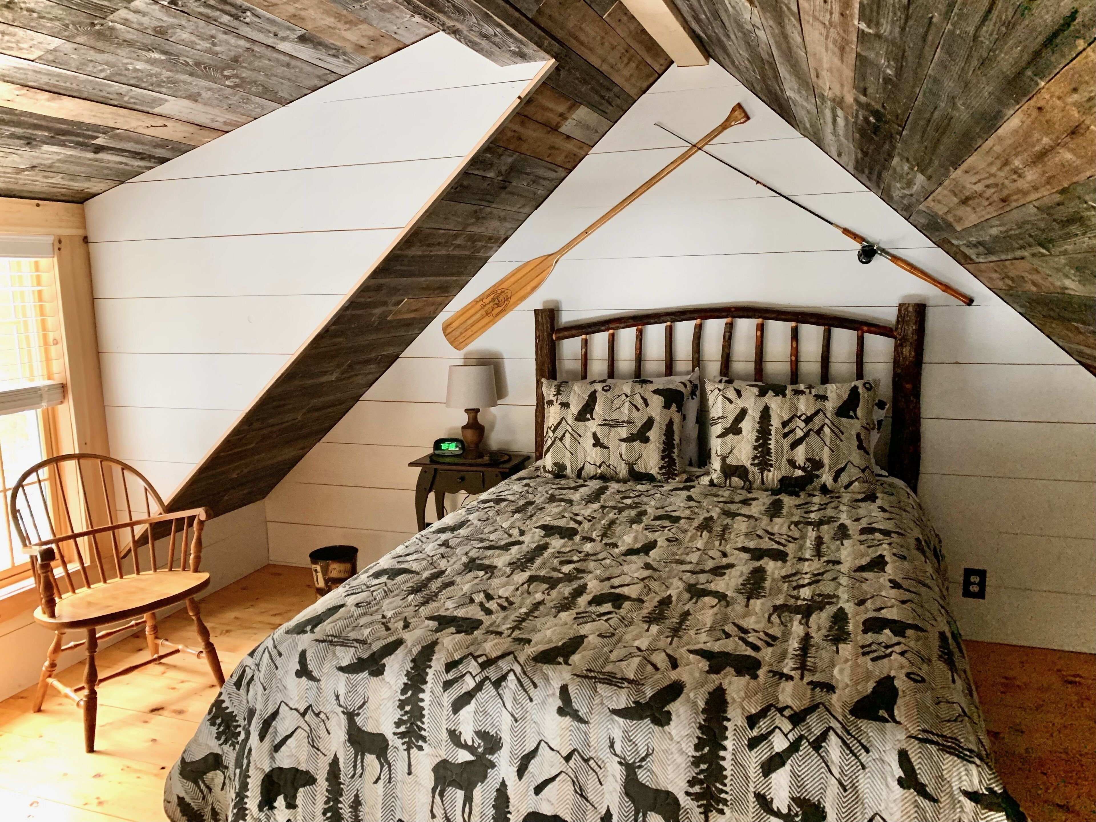 Et soveværelse med skråvæg, en seng med træramme, mønstret sengetøj, en stol, et sengebord med lampe og et vindue.