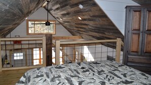 A bedroom with a sloped ceiling, wooden beams, a bed with patterned bedding, a window, and a wardrobe. - Brand new cabin in the Adirondacks near Whiteface Mountain! (Wilmington)