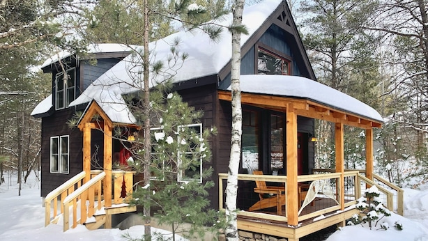 A cabin with a wooden porch and a stone foundation, surrounded by snow and trees. - Brand new cabin in the Adirondacks near Whiteface Mountain! (Wilmington)