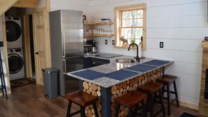 A compact kitchen with a built-in refrigerator, wooden bar stools, a sink, and a window with a view of trees. - Brand new cabin in the Adirondacks near Whiteface Mountain! (Wilmington)