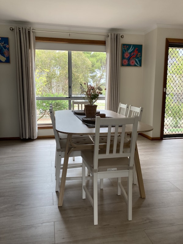 A dining area with a white table and chairs, a wicker centerpiece, and a view through a large window.