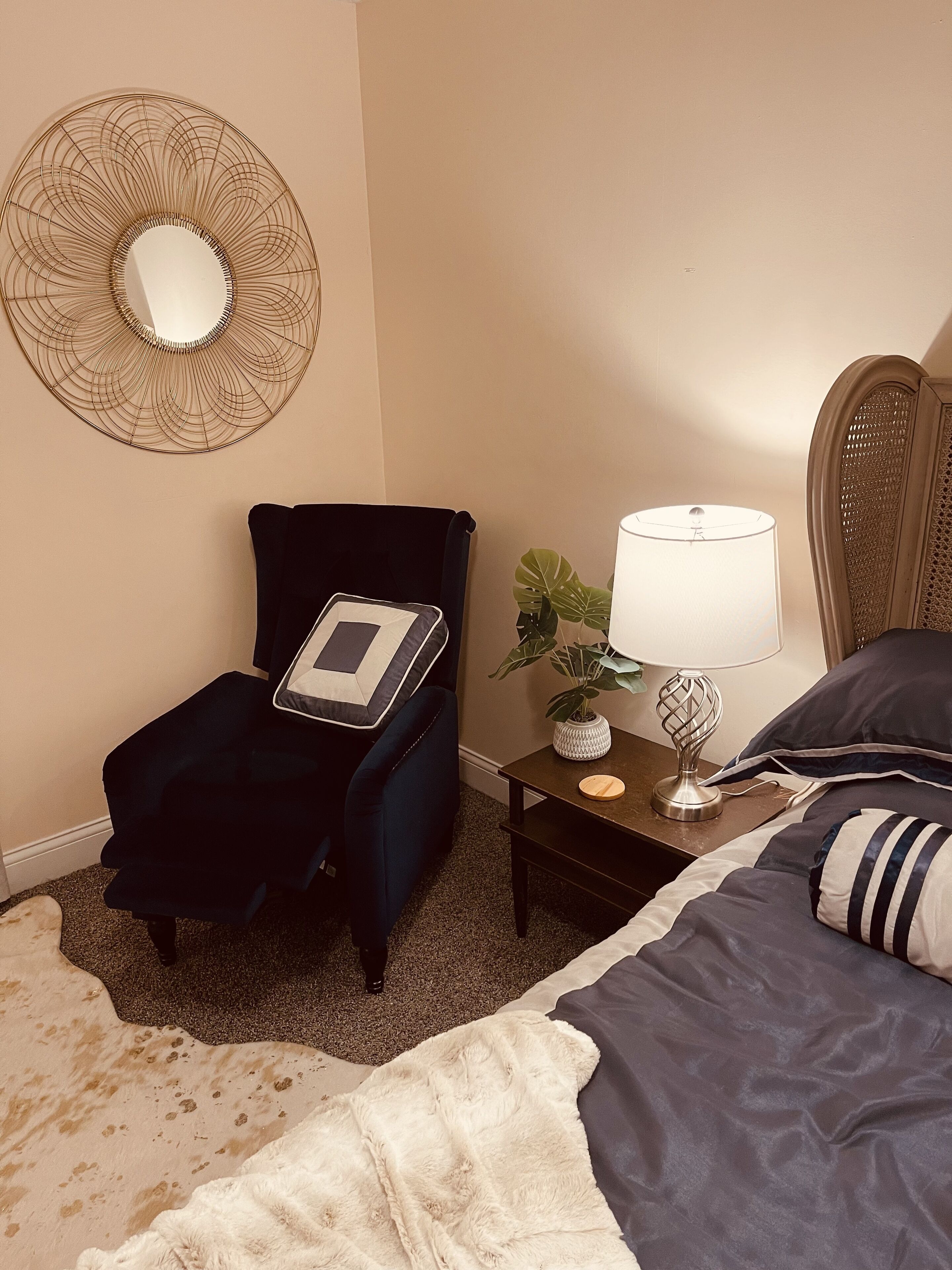 Una habitación de hotel con una cama, una silla con una computadora portátil, una mesita de noche con una lámpara y un espejo decorativo en la pared.