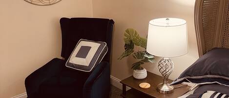Una habitación de hotel con una cama, una silla con una computadora portátil, una mesita de noche con una lámpara y un espejo decorativo en la pared.
