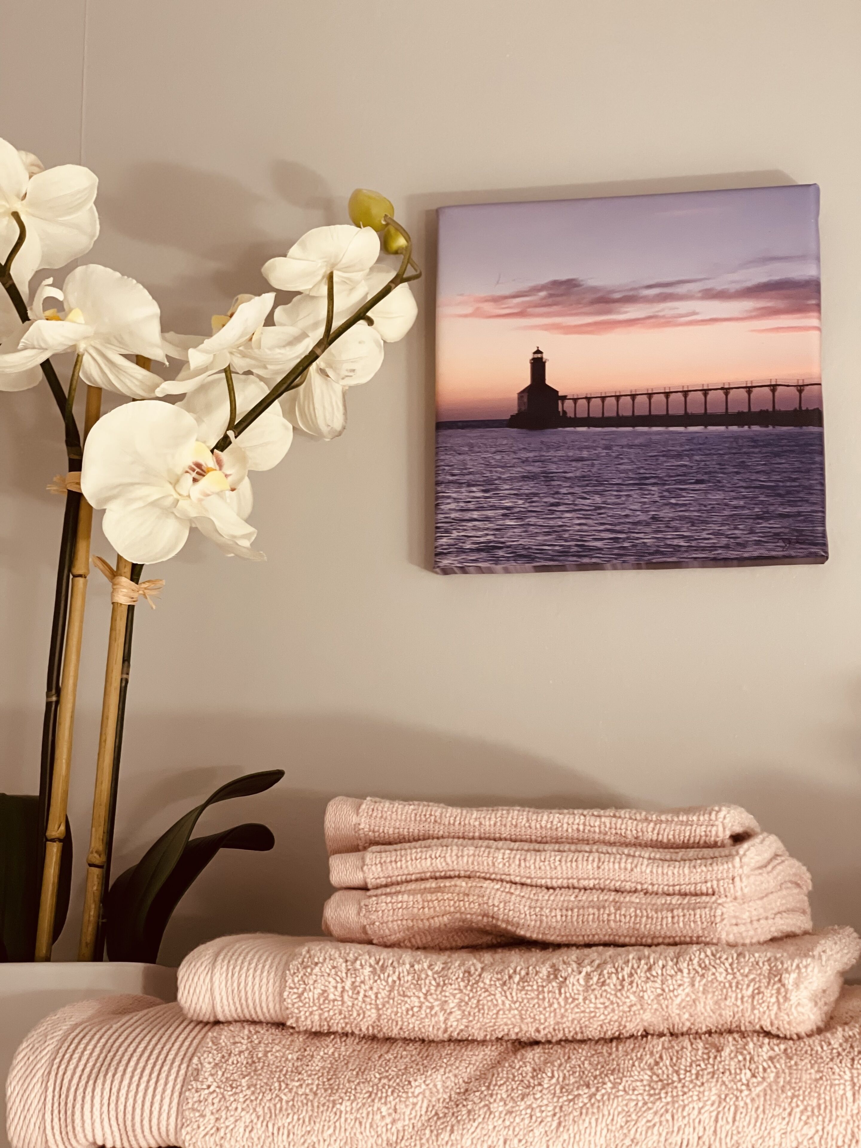 A room with a wall-mounted picture of a lighthouse at sunset, a bamboo plant, and a stack of pink towels.