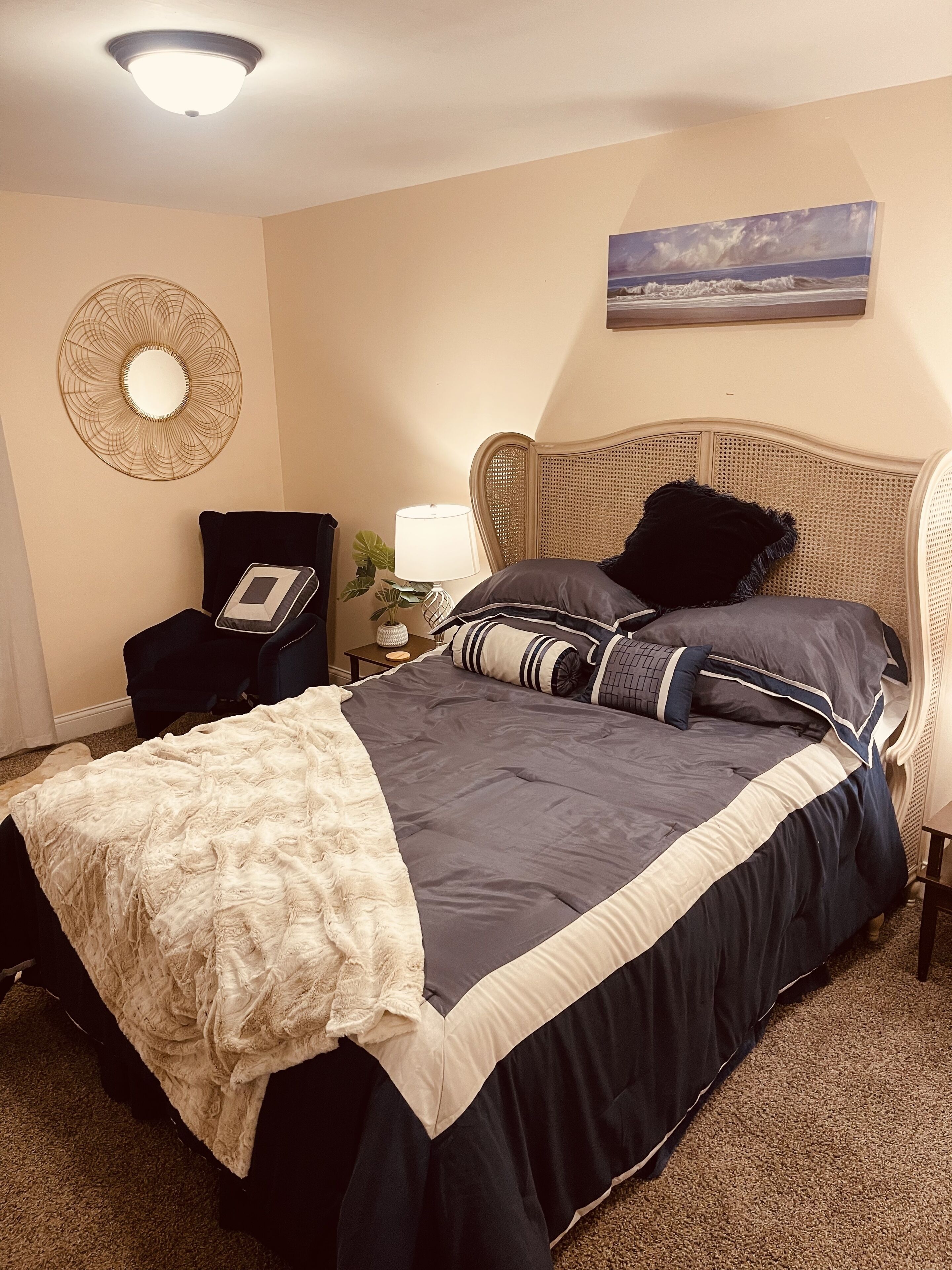 A neatly made bed with a dark blue and white duvet, a white throw blanket, and a rattan headboard. There is a bedside table with a lamp, a wall-mounted mirror, and a small potted plant.