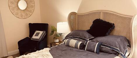 A neatly made bed with a dark blue and white duvet, a white throw blanket, and a rattan headboard. There is a bedside table with a lamp, a wall-mounted mirror, and a small potted plant.