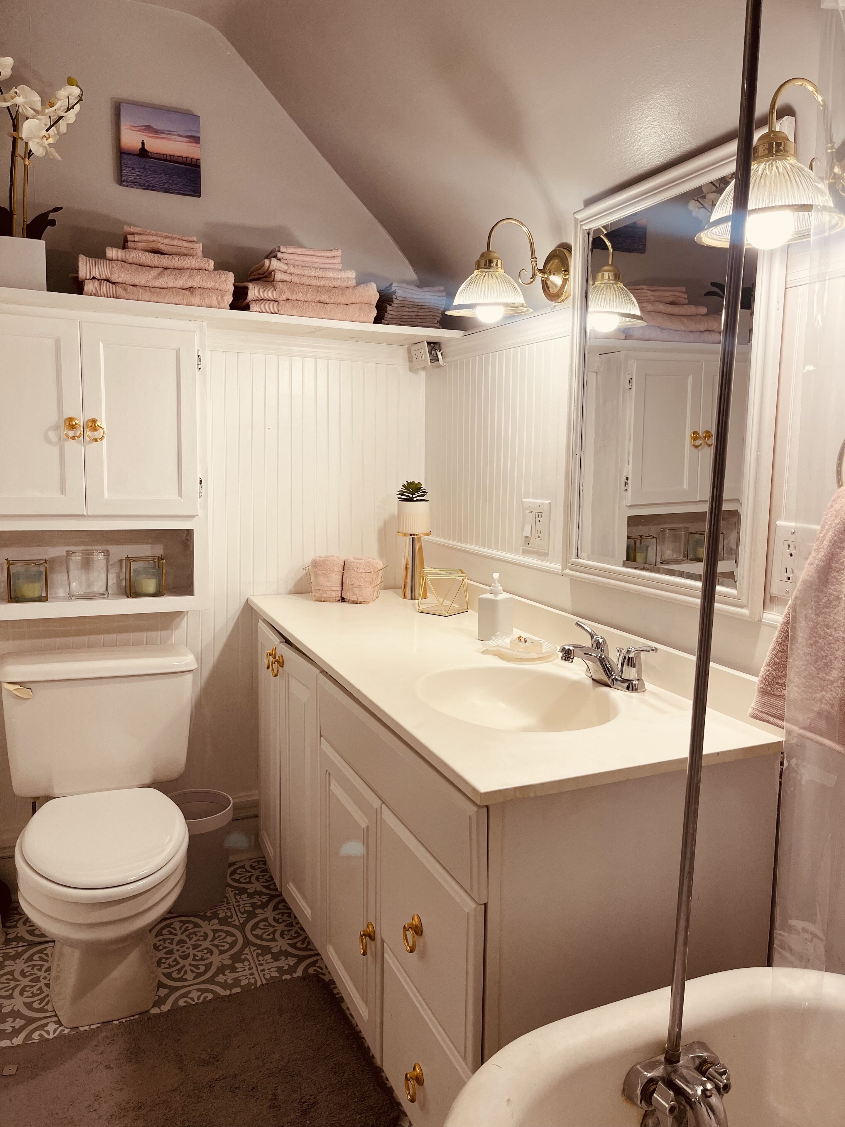 A bathroom with a white vanity, a sink, a toilet, and a bathtub. There are towels stacked on a shelf, a potted plant, and a mirror above the sink.