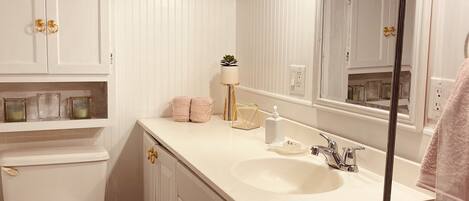 A bathroom with a white vanity, a sink, a toilet, and a bathtub. There are towels stacked on a shelf, a potted plant, and a mirror above the sink.
