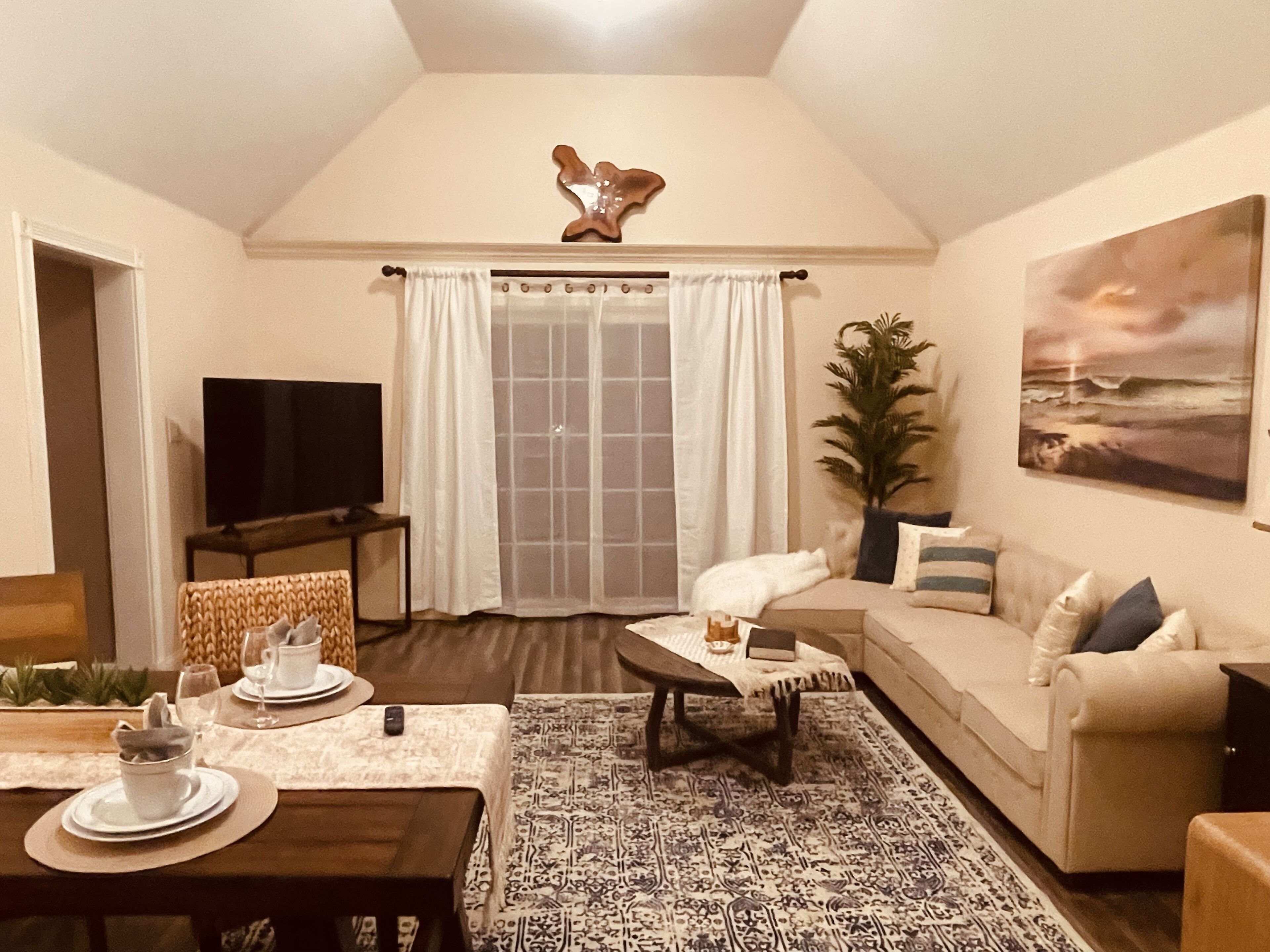 A cozy living room with a sofa, a coffee table, and a TV. There's a window with curtains and a plant on the right.
