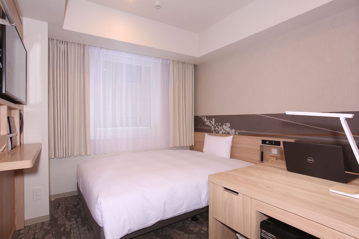 A hotel room with a bed, a desk with a laptop, a TV, and a window with curtains.