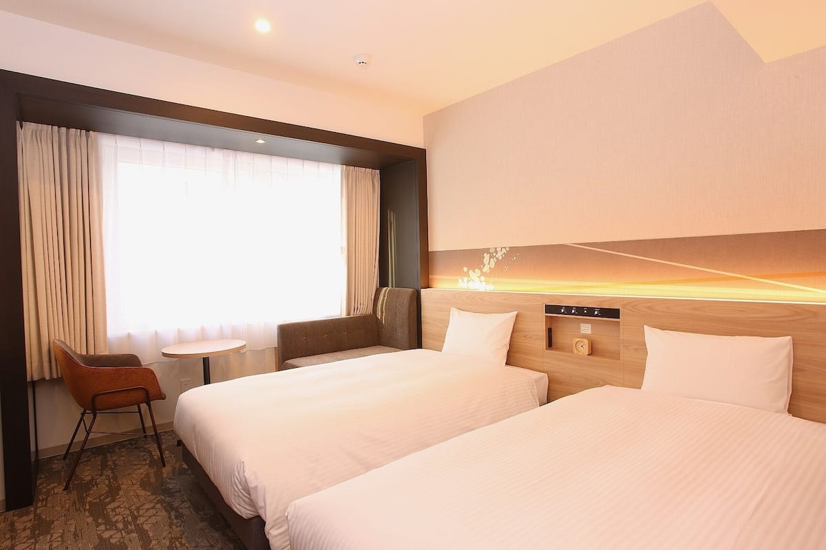A hotel room with two beds, a small table, a chair, and a view of the sunset through the window.