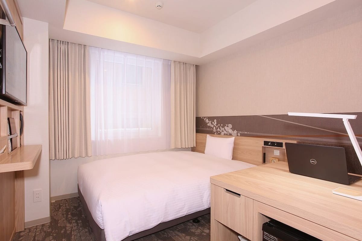 A hotel room with a bed, a desk with a laptop, a TV, and a window with curtains.