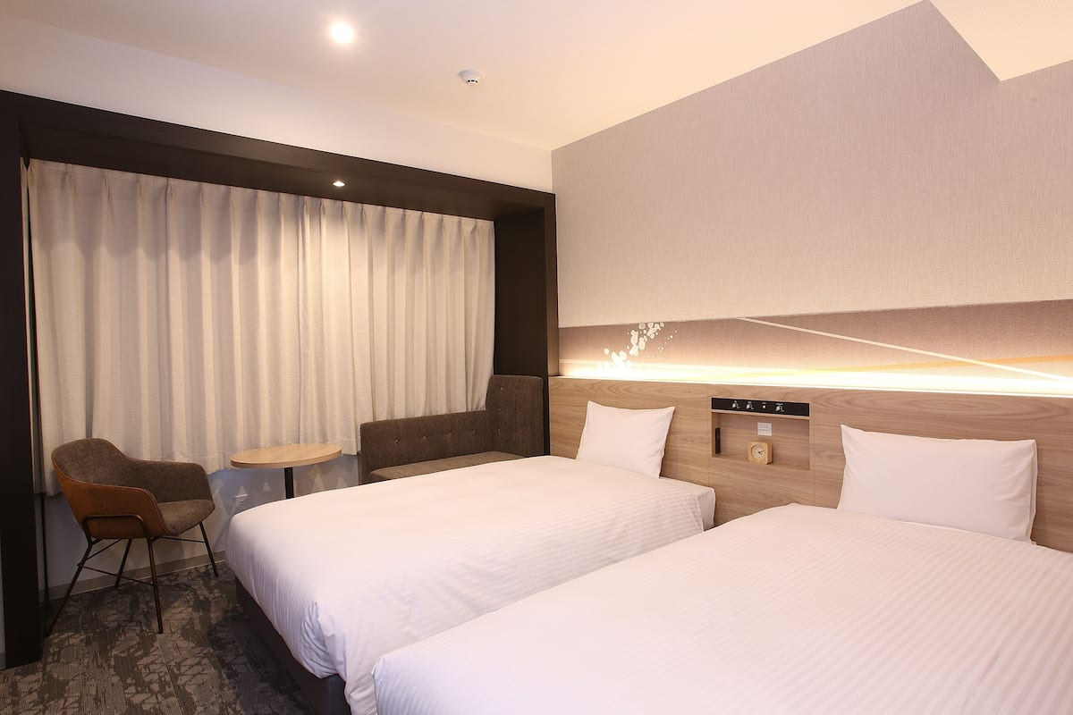 A hotel room with two beds, a small table, a chair, and a clock on the wall.