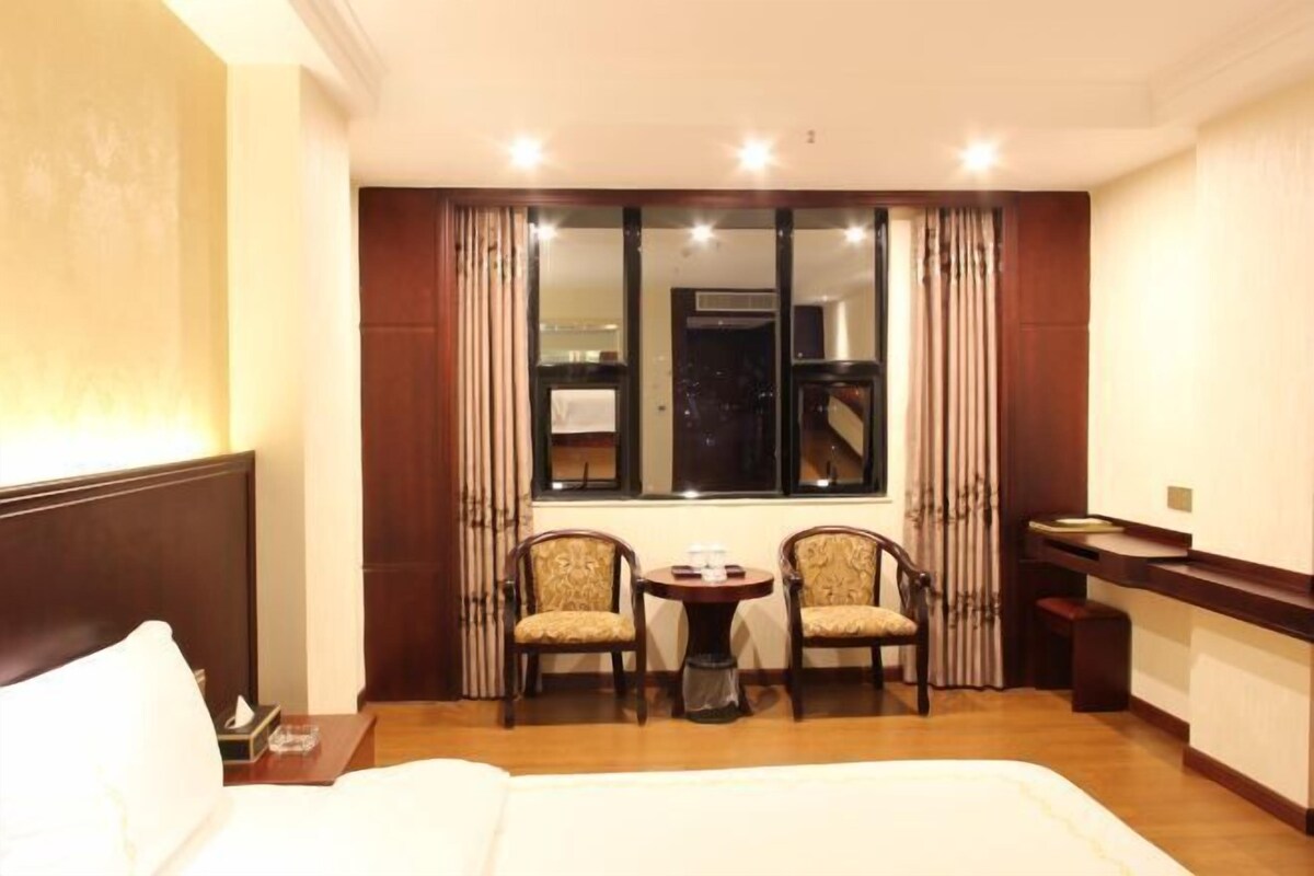 A hotel room with a bed, two chairs, a small table, a window with curtains, and a view of a building outside.