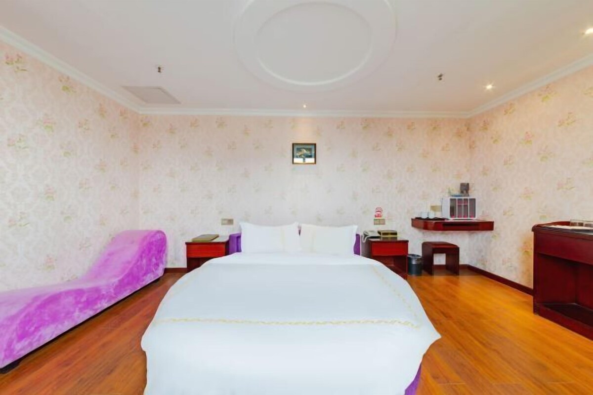 A hotel room with a large bed, a purple sofa, a wooden floor, and a small table with a lamp.
