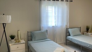 A bedroom with two single beds, a bedside table with a lamp, and a window with sheer curtains. - A Great Boca Home Stay (Boca Raton)