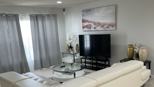 A modern living room with a white leather sofa, a glass coffee table, a flat-screen TV, and decorative vases. - A Great Boca Home Stay (Boca Raton)