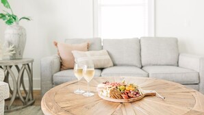 A round wooden table with a charcuterie board and two glasses of white wine, set in a living room with a grey sofa and a vase of flowers. - The Sand Haus - Steps to Beach - 7 Separate Beds - Pet Friendly - Pool - Grill (Gulf Shores)