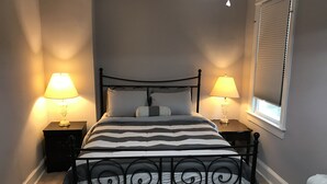 A bedroom with a bed, two bedside tables with lamps, a window with blinds, and a radiator. - Brookland Quiet Living (Washington)