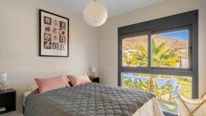 Quarto com cama grande, quadro na parede, luminária pendente e janela com vista para palmeiras e colinas.