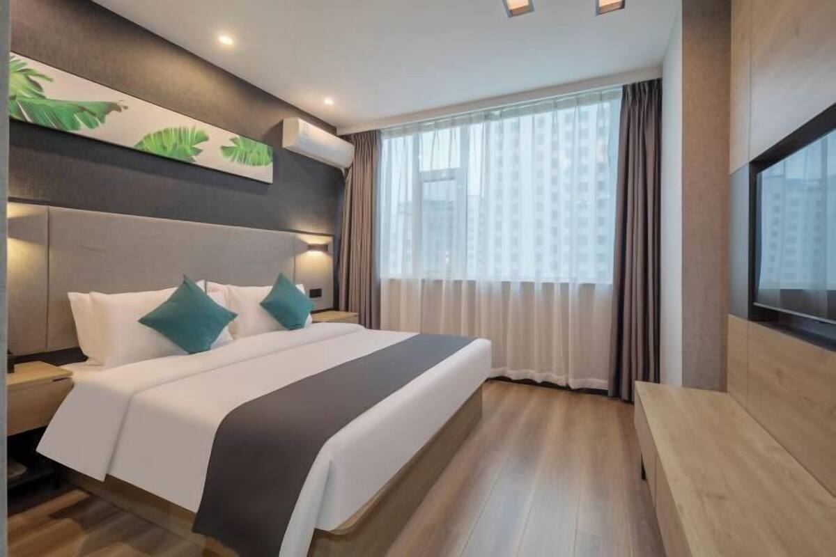 Una habitación de hotel moderna con una cama grande, una mesita de noche, un banco y un ventanal con cortinas.
