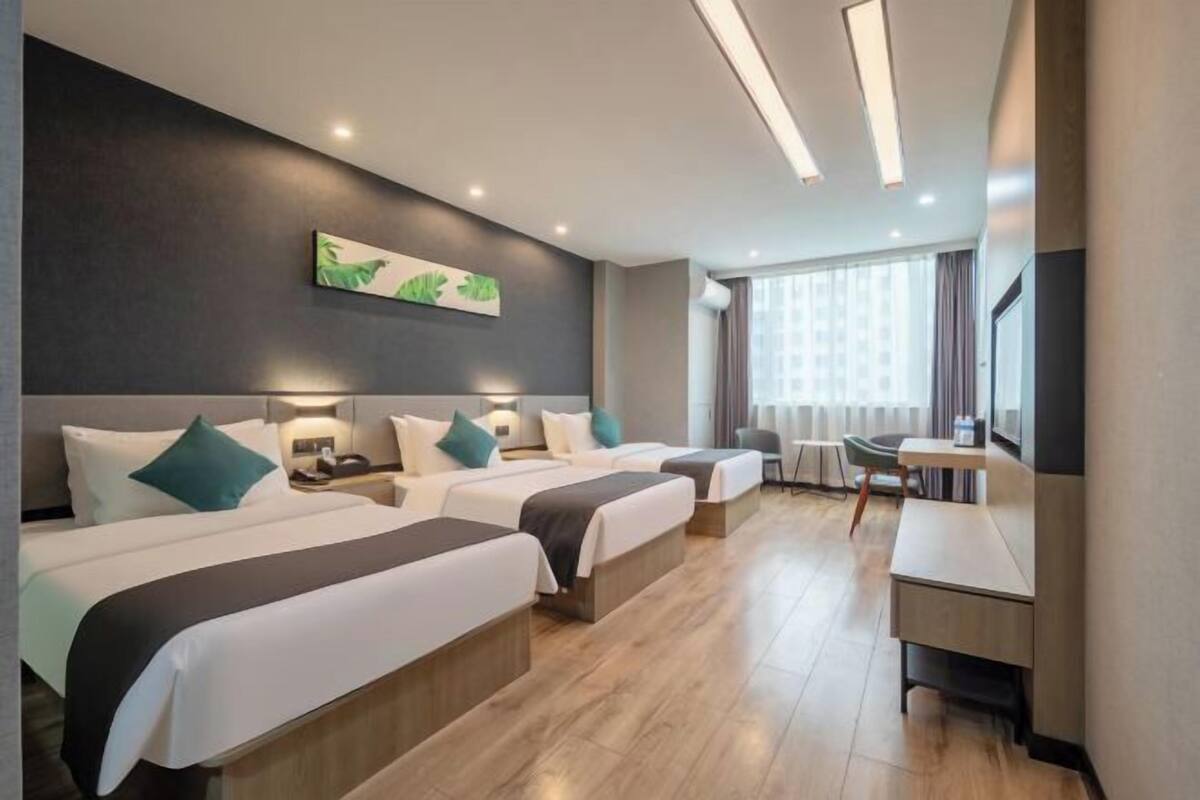 Habitación de hotel con tres camas, un escritorio, una silla y un ventanal con cortinas.