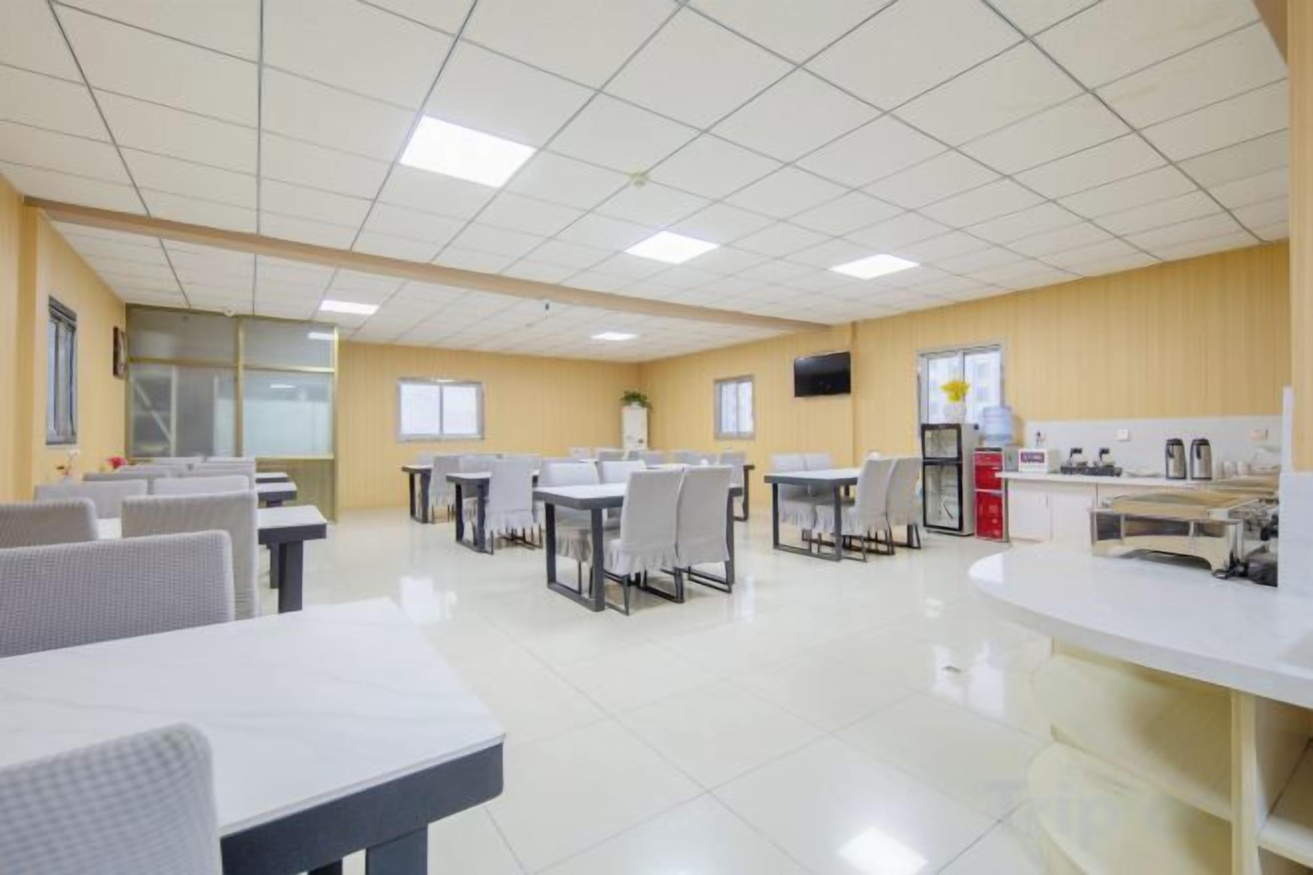 Un comedor limpio y bien iluminado con varias mesas y sillas, una encimera de cocina y un televisor montado en la pared.