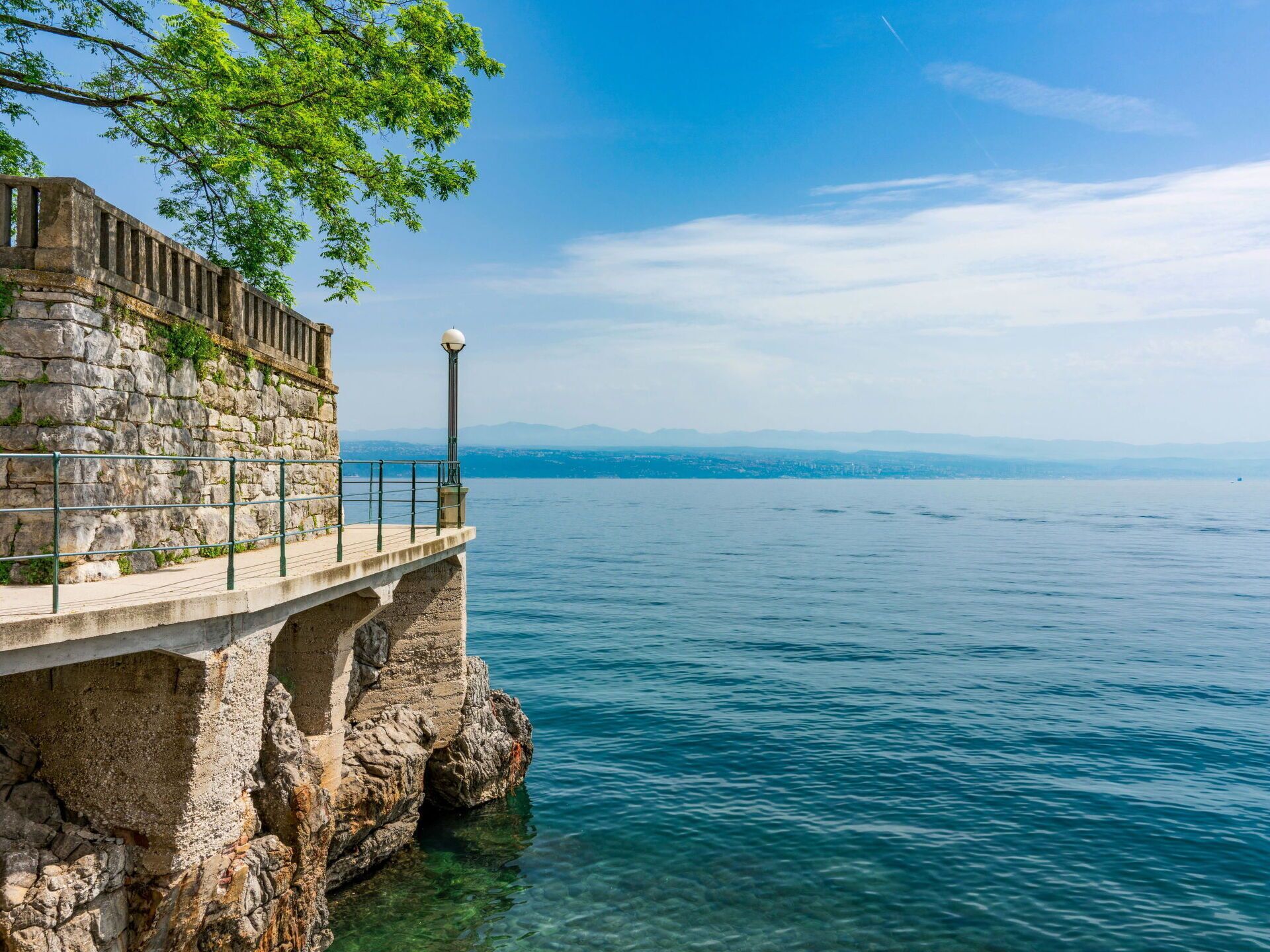 Un muelle de piedra se adentra en una masa de agua tranquila, con vistas despejadas a montañas distantes y al perfil urbano de una ciudad.