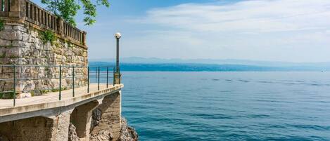 Un muelle de piedra se adentra en una masa de agua tranquila, con vistas despejadas a montañas distantes y al perfil urbano de una ciudad.