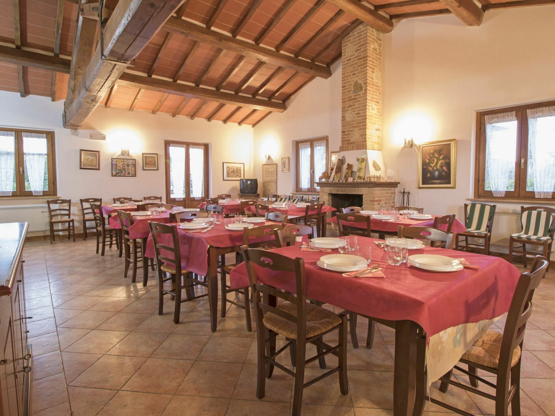 A dining hall with multiple tables set for meals, wooden chairs, a fireplace, and framed artwork on the walls.