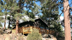 A cabin with a wooden deck and a large window, surrounded by trees and rocky terrain. - 2 Bedroom with Great View from Wrap-Around Deck (Estes Park)