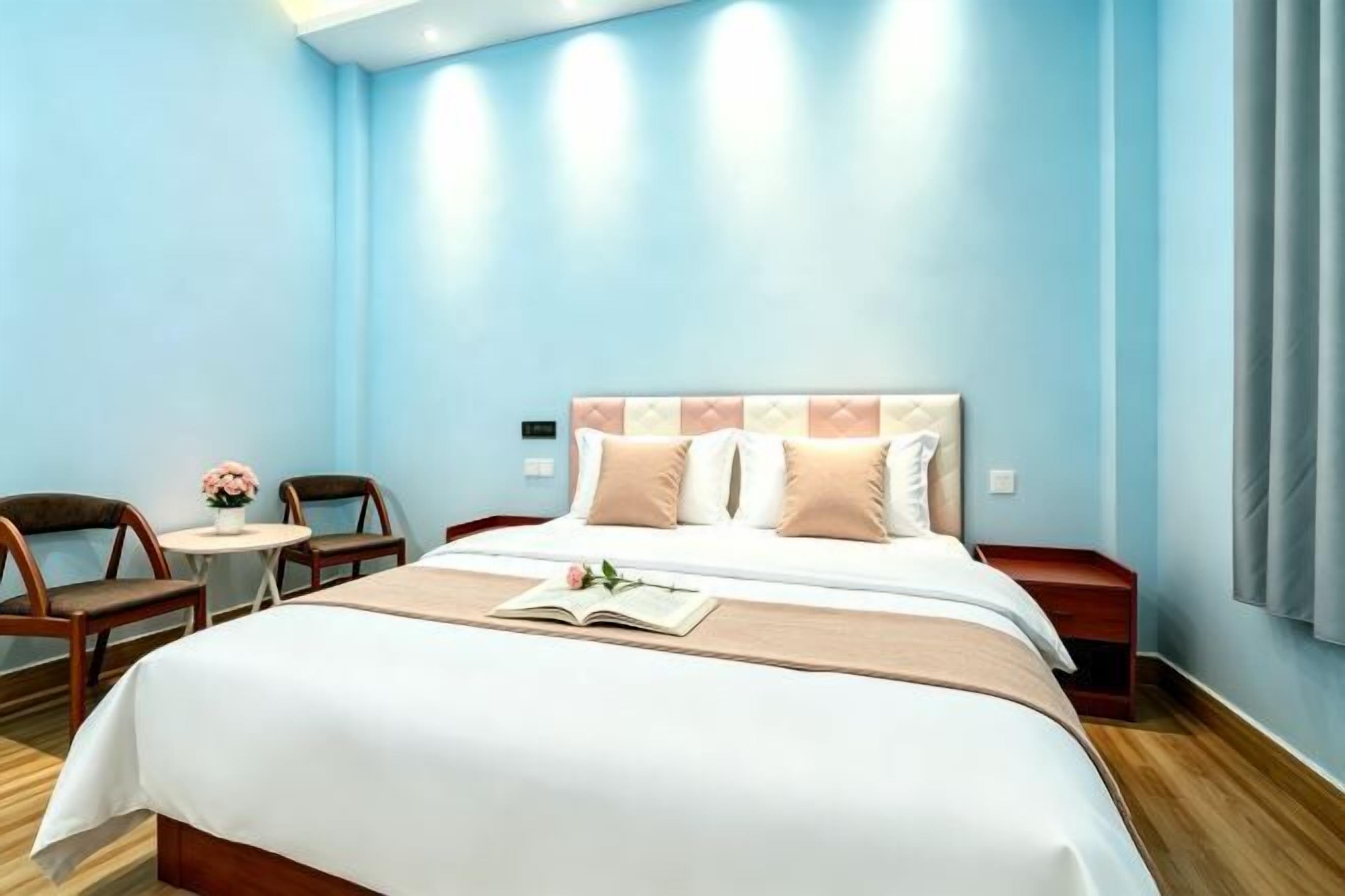 A hotel room with a large bed, a bedside table, a chair, a small round table, and a potted plant.