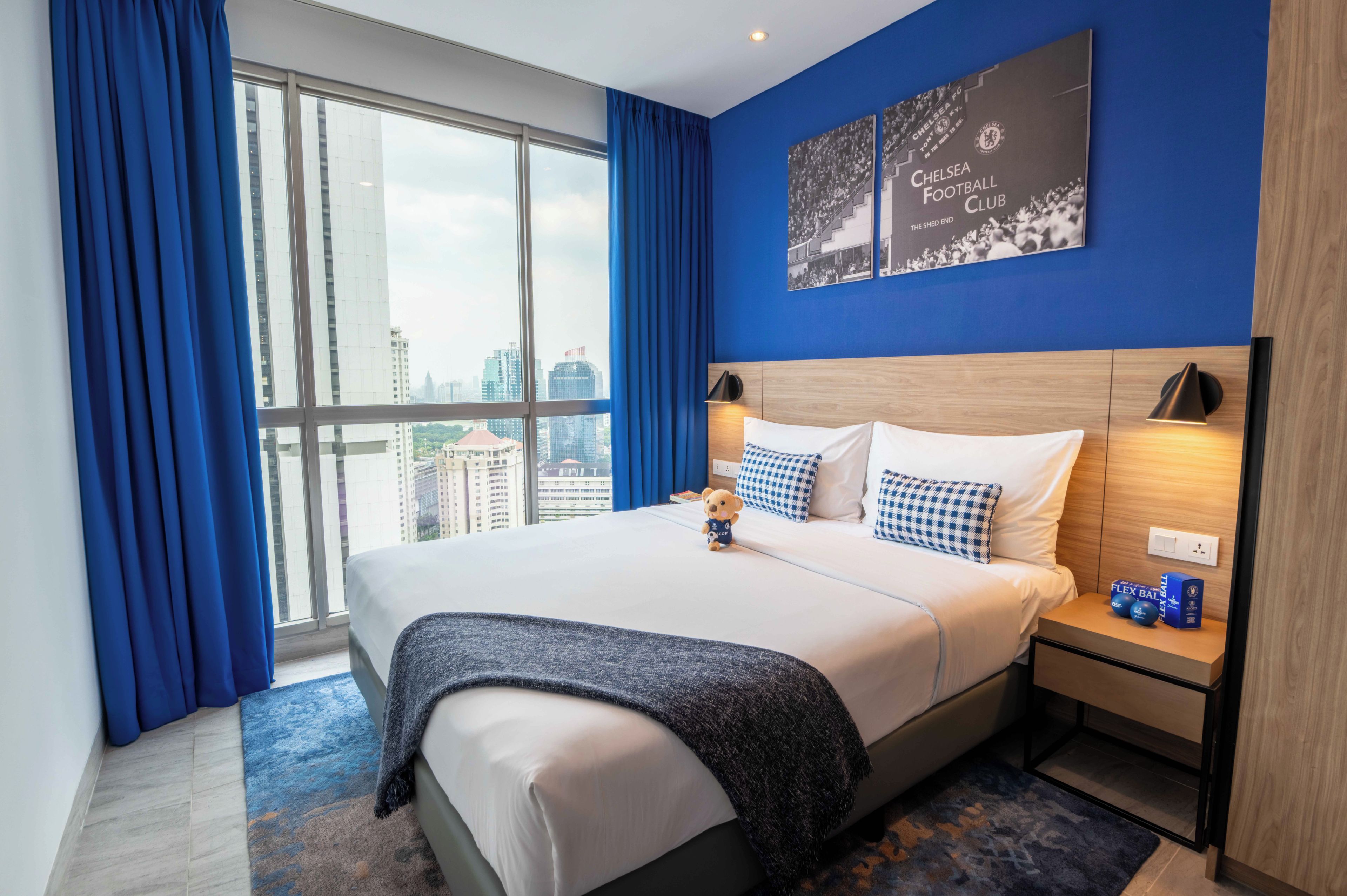 Une chambre d’hôtel avec un grand lit, une table de chevet, un ours en peluche, une vue sur la ville et des objets de collection du Chelsea Football Club.