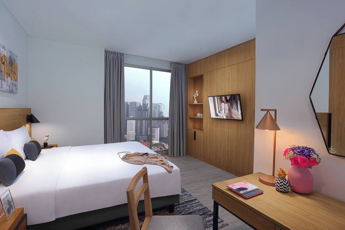 Une chambre d’hôtel moderne avec un grand lit, un bureau, une chaise, une lampe de bureau, un vase de fleurs, un cadre photo et une vue sur la ville par une grande fenêtre.