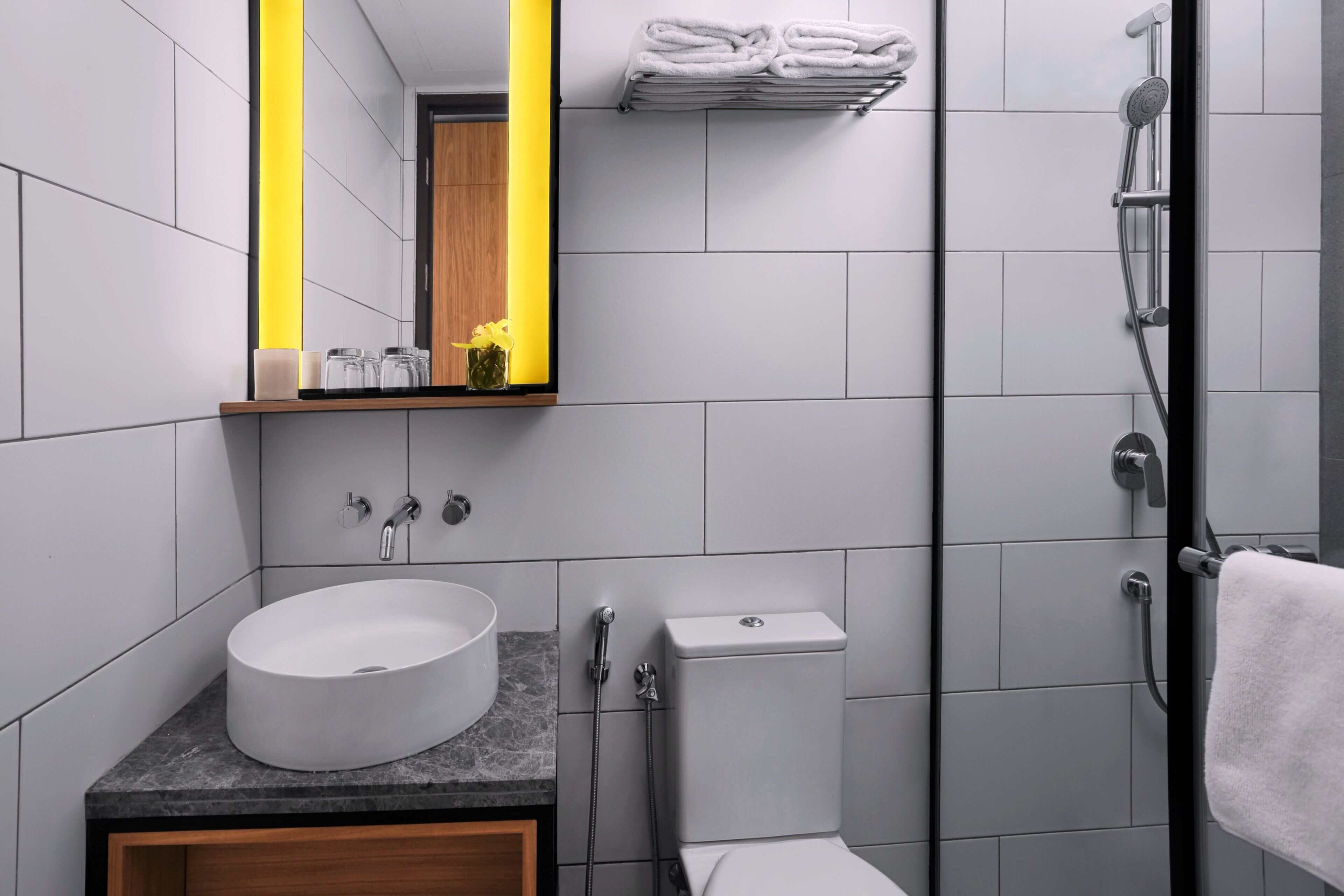 A modern bathroom with a round sink, a toilet, a shower, and a mirror with a yellow light strip.