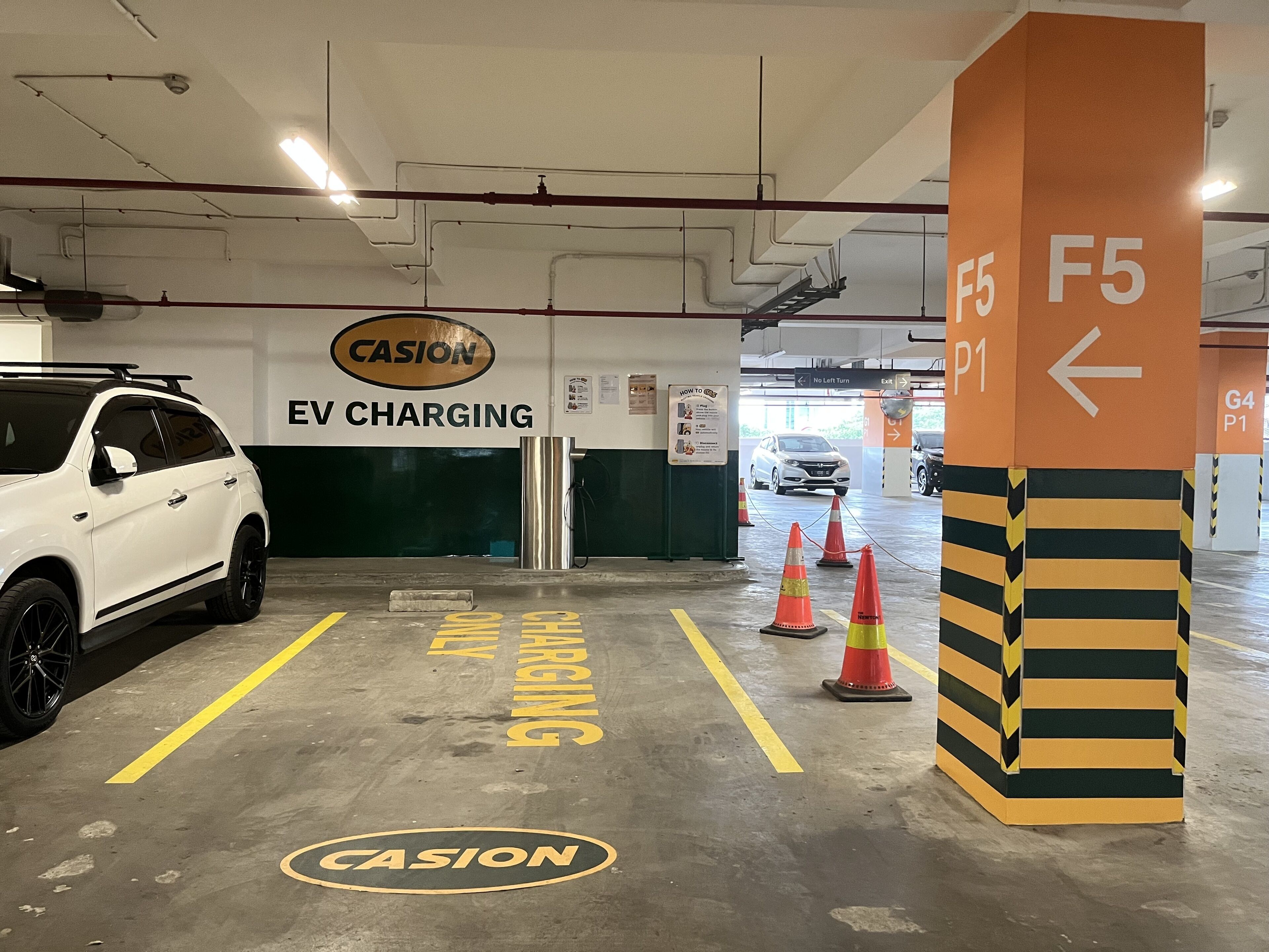 An EV charging station area with a white SUV parked, directional signs, and a Casion logo on the floor.
