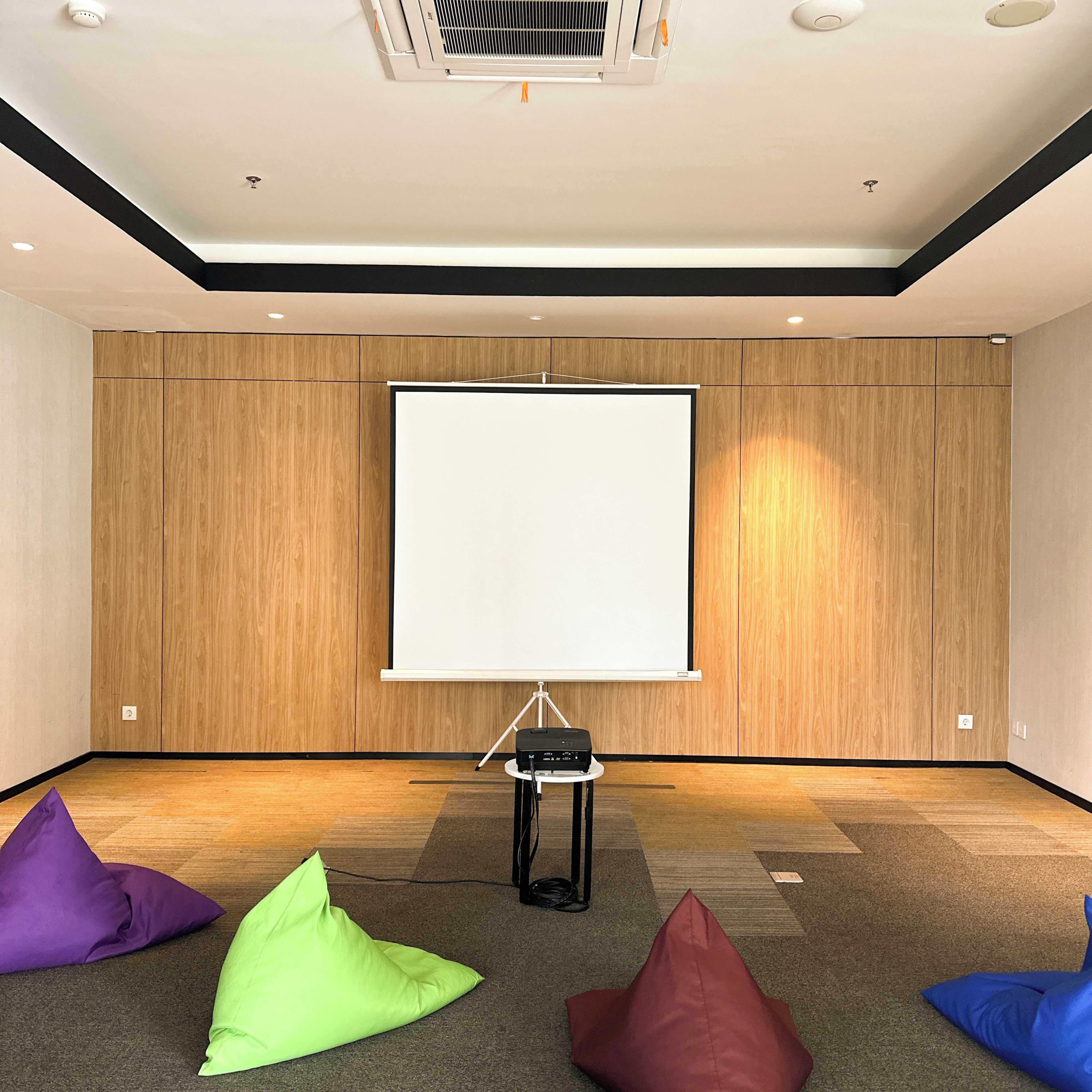 Un salón de conferencias con una gran pantalla de proyección, paredes de paneles de madera y coloridos puffs en el suelo.