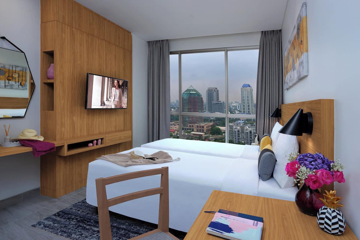 Une chambre d’hôtel comprenant un lit, un bureau, une chaise, une télévision, une fenêtre donnant sur la ville et une armoire en bois.