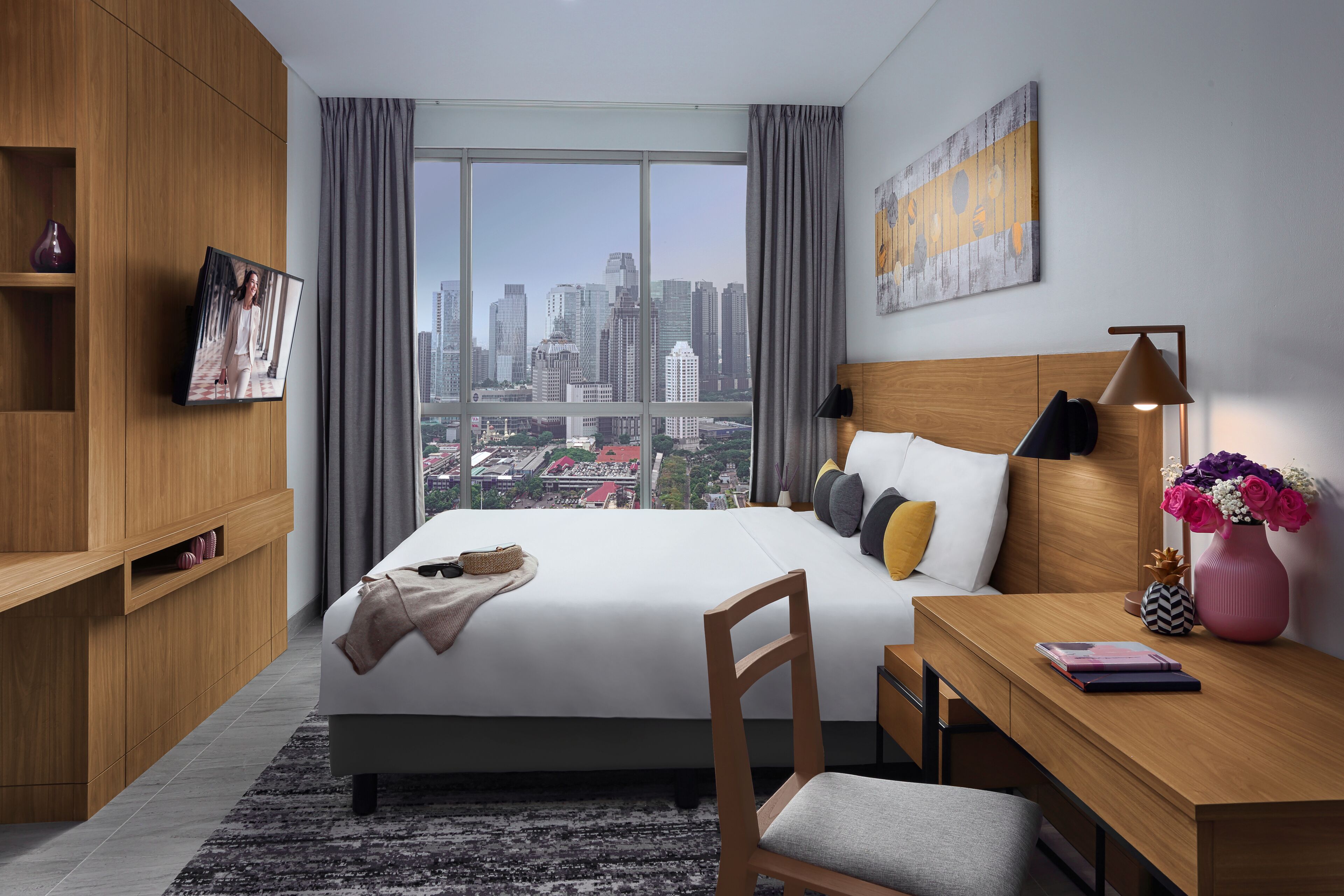 Una habitación de hotel moderna con una cama grande, un escritorio, una silla, un televisor y vistas a la ciudad.