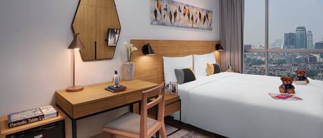 A hotel room with a wooden desk, a bed with white bedding, a city view, and a painting on the wall.