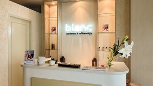 Un comptoir d’accueil avec une pancarte indiquant « blanc wellness & réflexologie ». On y trouve des étagères remplies de produits, un vase avec des fleurs et un magazine posé sur le comptoir.