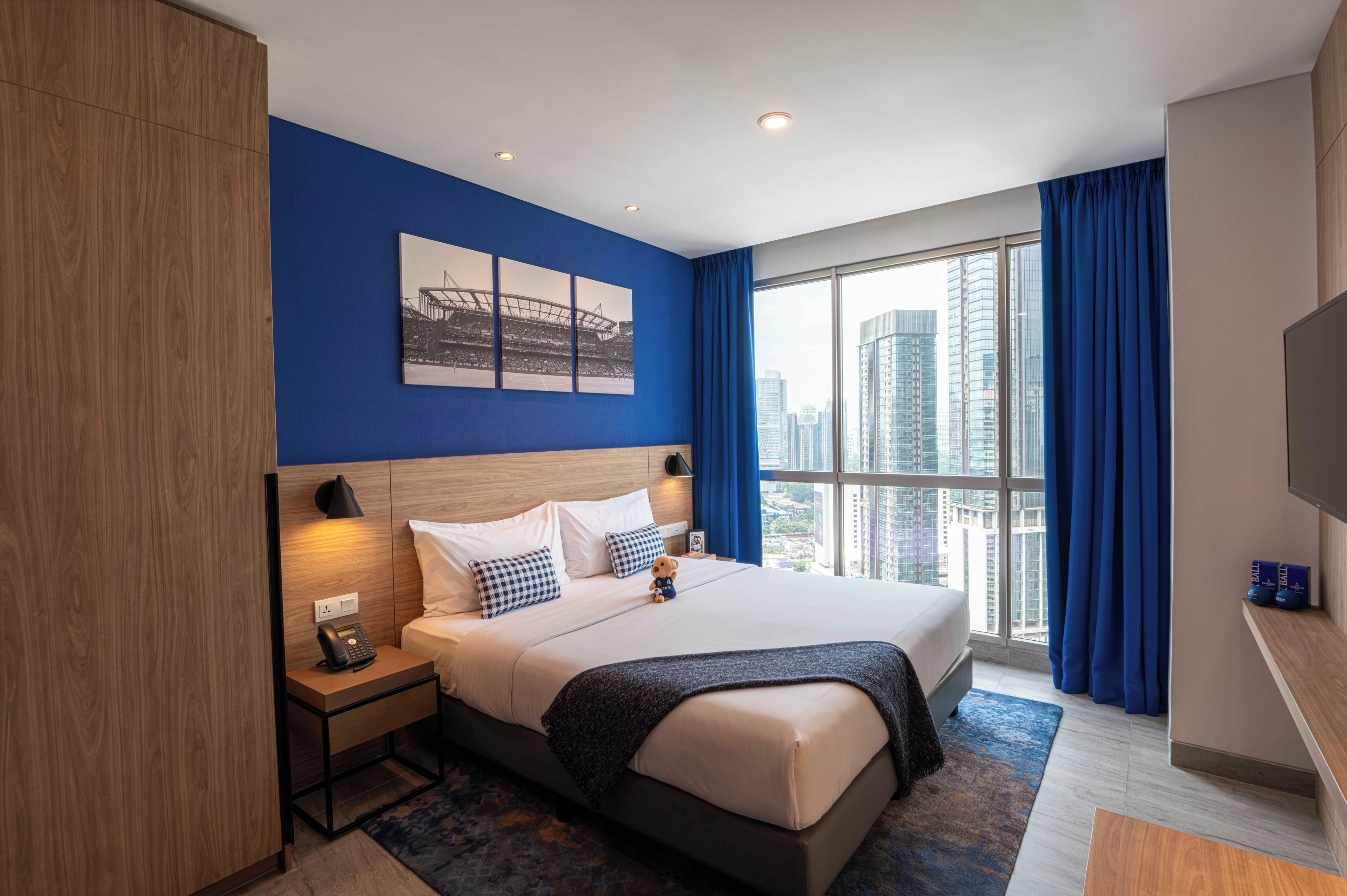 A hotel room with a large bed, a nightstand, a telephone, a painting on the wall, a window with curtains, and a view of the city.