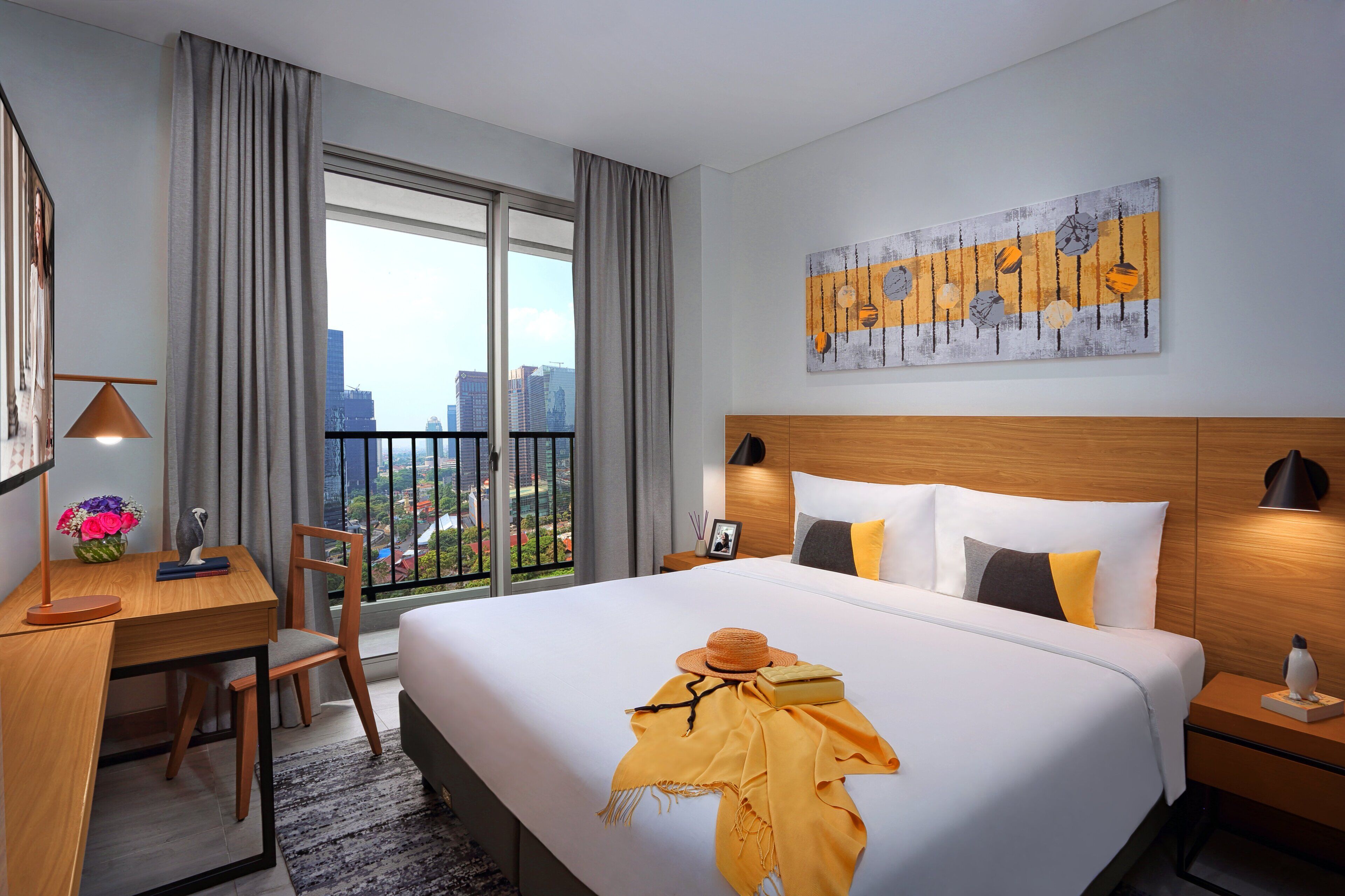 A hotel room with a large bed, a desk with a lamp, a chair, and a view of the city through a balcony.