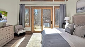 Un dormitorio con una cama grande, televisión, escritorio y una silla. Tiene un balcón con vistas a árboles y una terraza de madera.