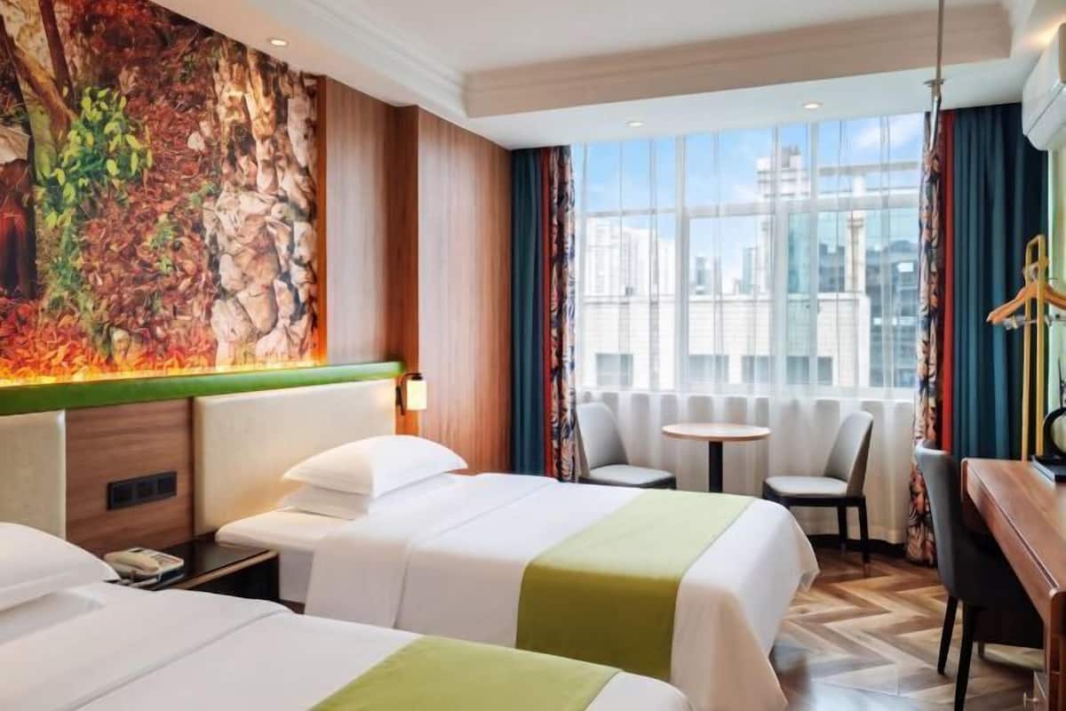A hotel room with two beds, a large painting, a city view, a small table, and a desk.