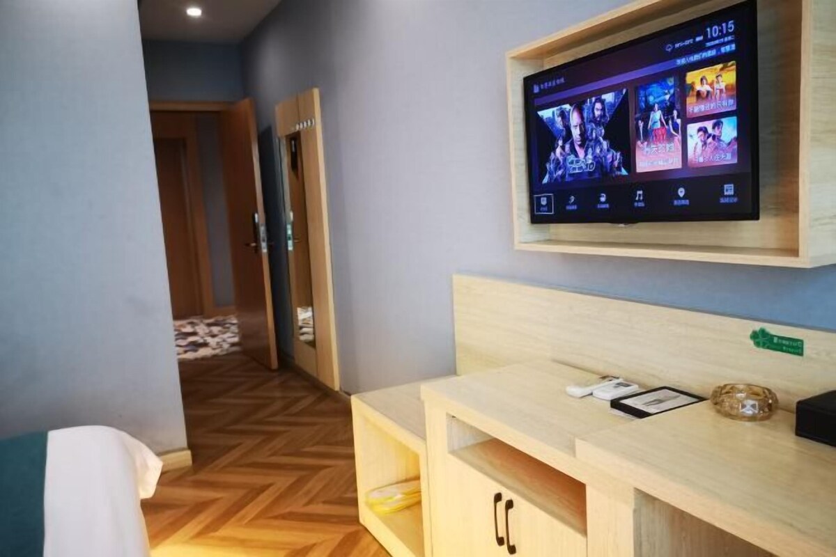 A hotel room with a wooden floor, a bed, a TV mounted on the wall, and a wooden desk with a nameplate.