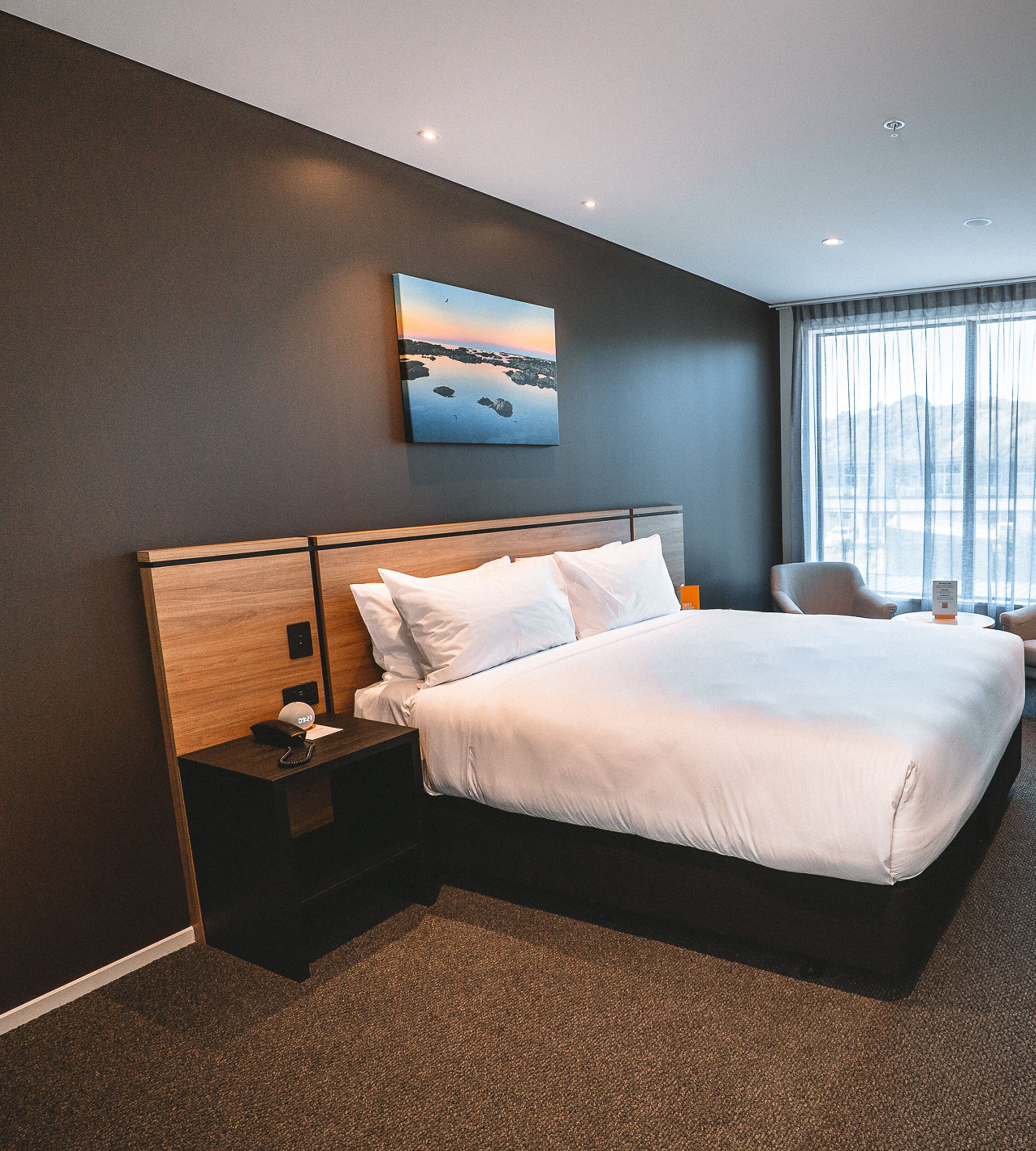 A modern hotel room with a large bed, a nightstand, a chair, and a painting on the wall.