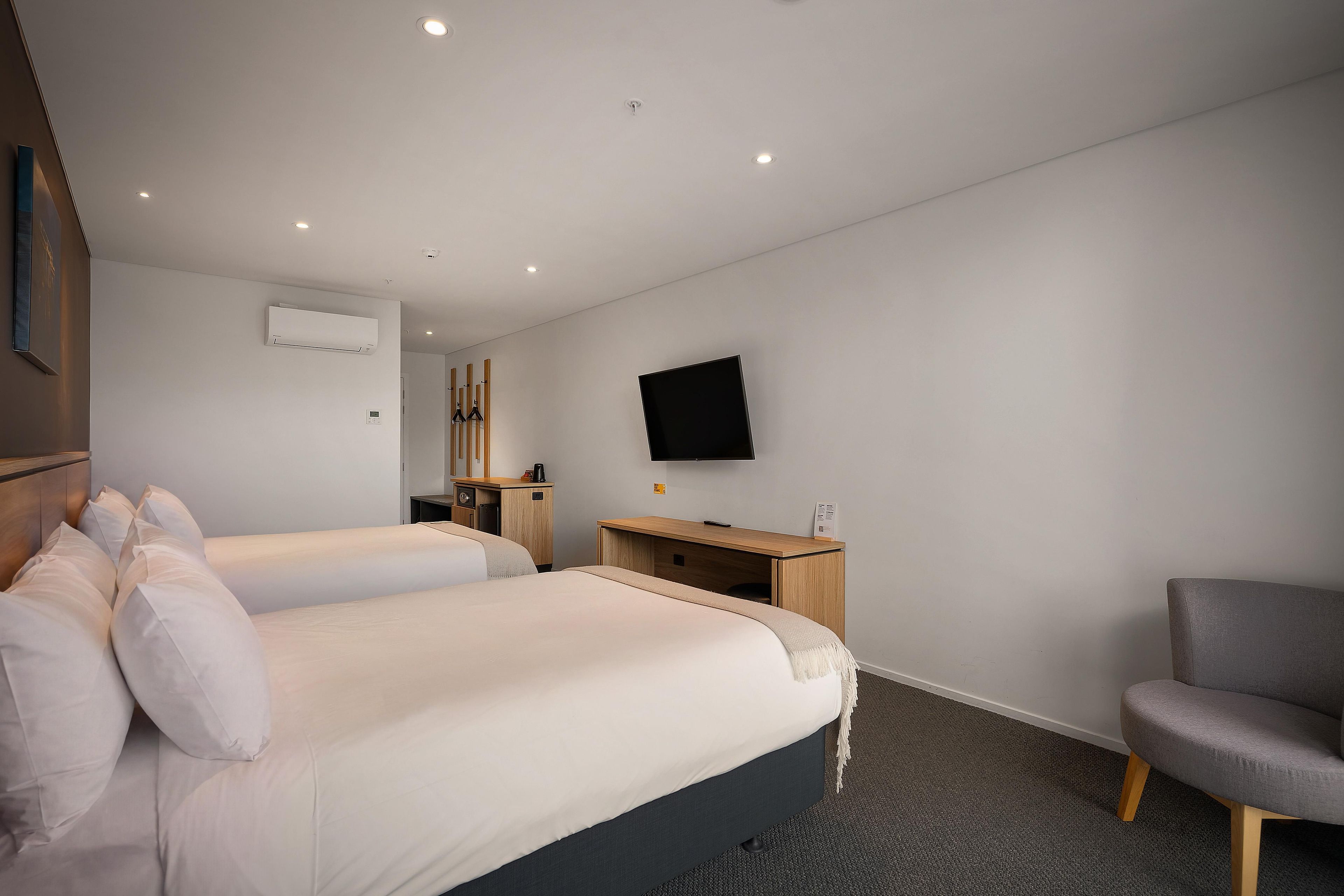 A hotel room with two beds, a desk, a chair, a flat-screen TV, and an air conditioning unit.