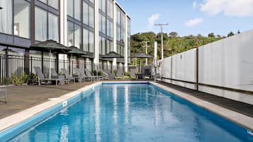 A swimming pool with lounge chairs and umbrellas, a building with large windows, and a view of a hilly landscape.
