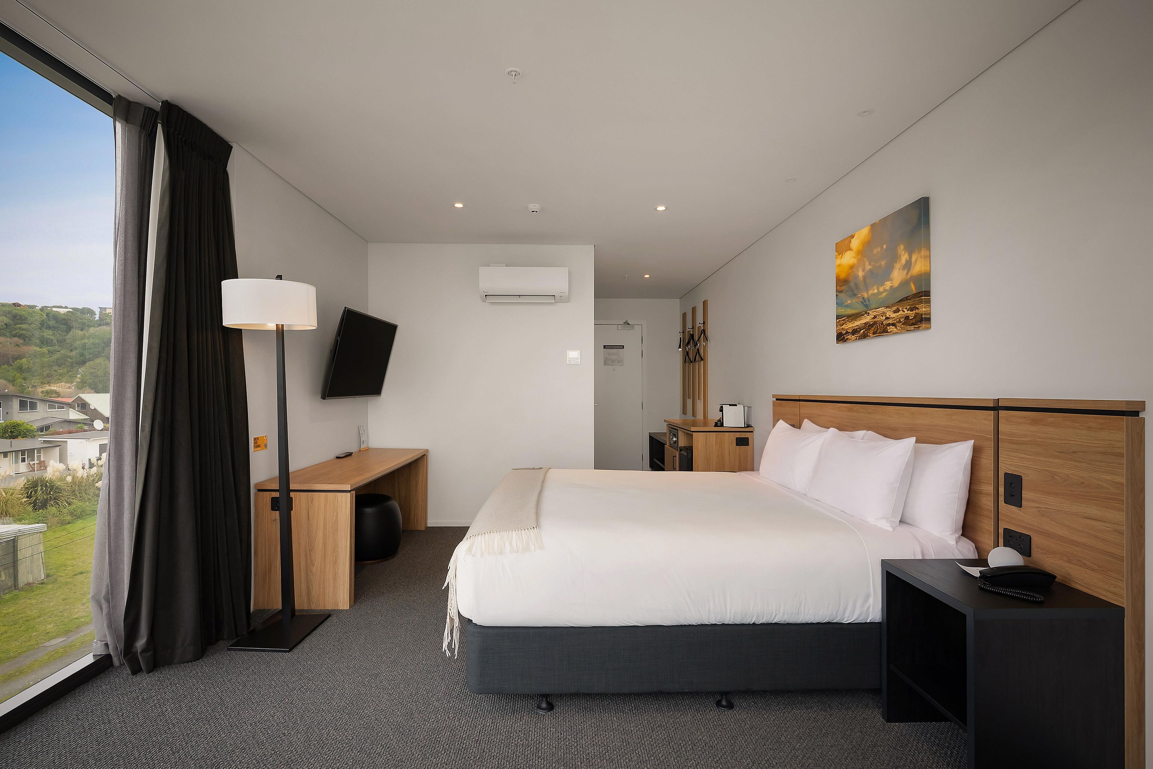 A hotel room with a bed, bedside table, lamp, TV, air conditioning unit, and a painting on the wall.